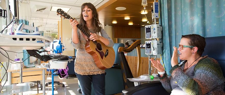 An SPU music therapy alum plays her guitar while a patient claps.