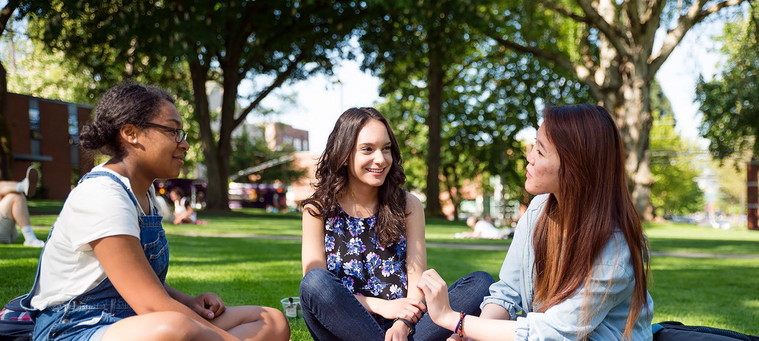 Three students talking while sitting on the grass in Tiffany Loop/